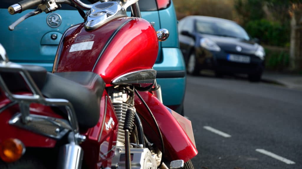 A red motorbike parked on a residential street with a blue car in the background