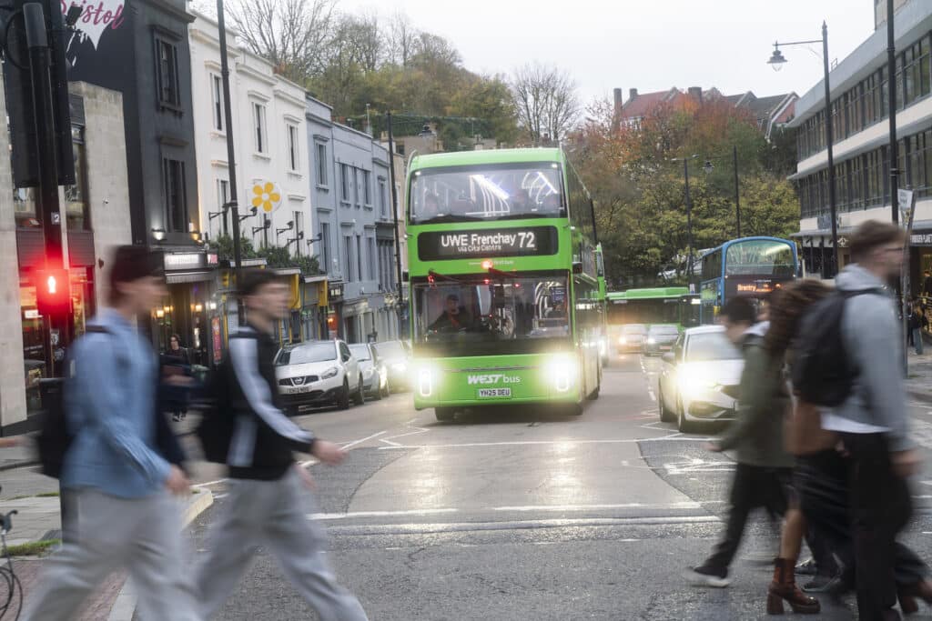 A green WEST bus traveling in Clifton Triangle with other buses stopped at a bus stop in the background, in the foreground are pedestrians crossing the road