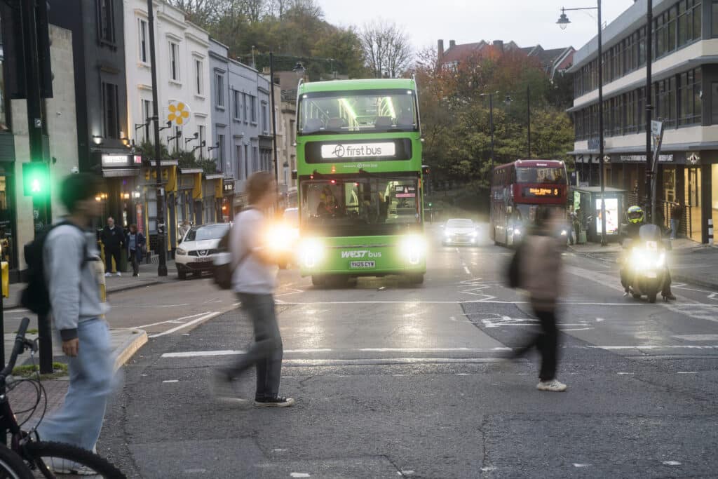 A green WEST bus traveling in Clifton Triangle with a Red bus stopped at a bus stop in the background, in the foreground are pedestrians crossing the road