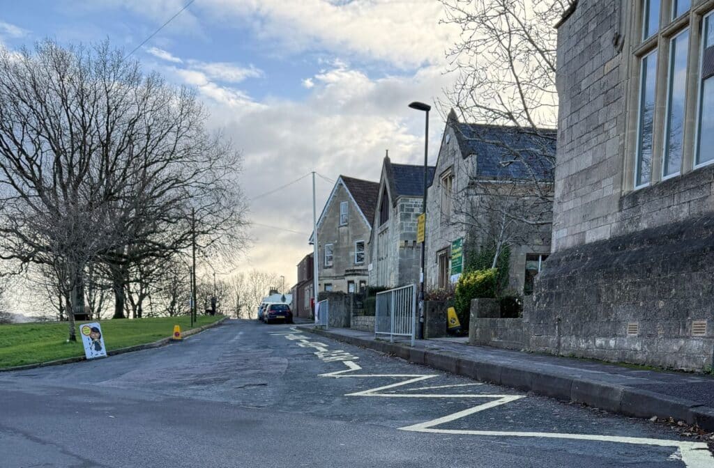 Residential street outside stone buildings with school zigzag road markings, parked cars, and a grassy area with trees under a partly cloudy sky.