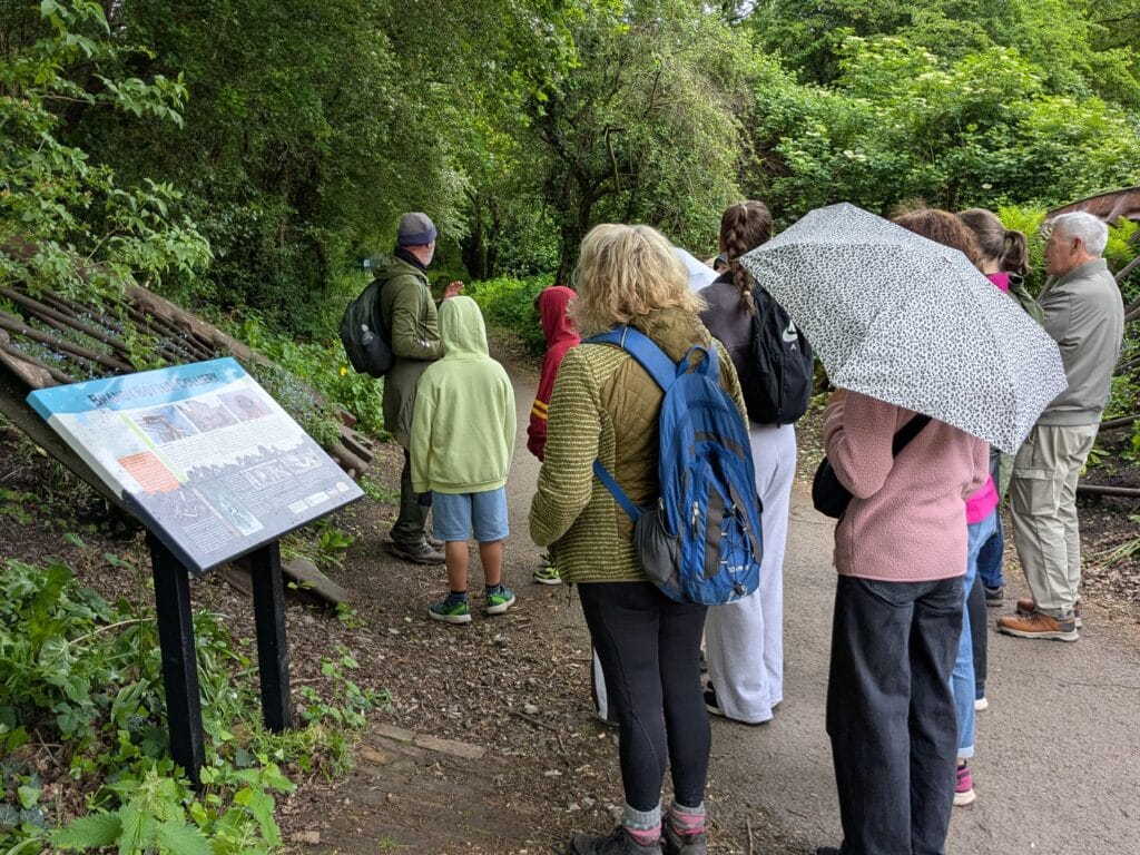 Photo of guided walk group on a woodland path at Brandy Bottom historic colliery, listening to a speaker beside an information board about the site.