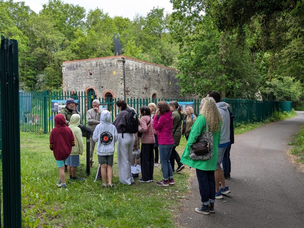Photo of guided walk group gathered outside the historic Brandy Bottom colliery, listening to a speaker beside the fenced stone building and woodland path.