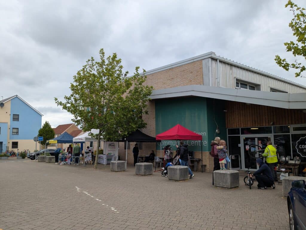 Photo of sustainable travel event outside a community centre with several pop-up gazebos, people gathered, and activities including a bike repair stand and mechanic and information stalls.