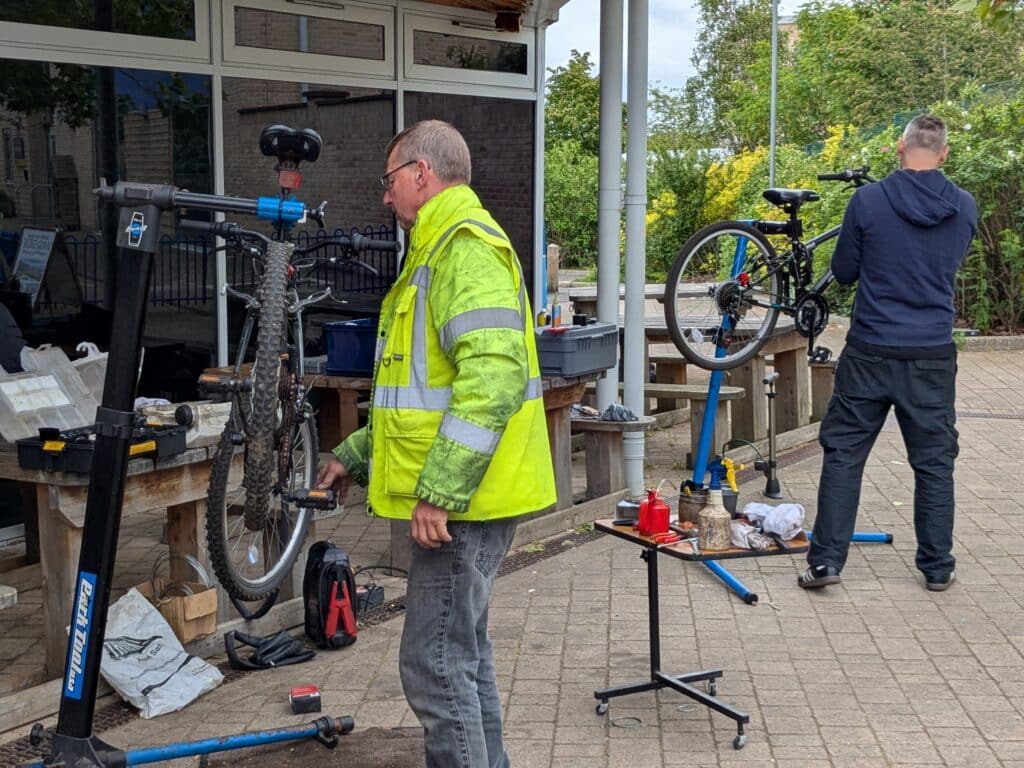 Photo of outdoor bike repair area with two people servicing bicycles on stands, with tools and equipment set up outside the community centre.