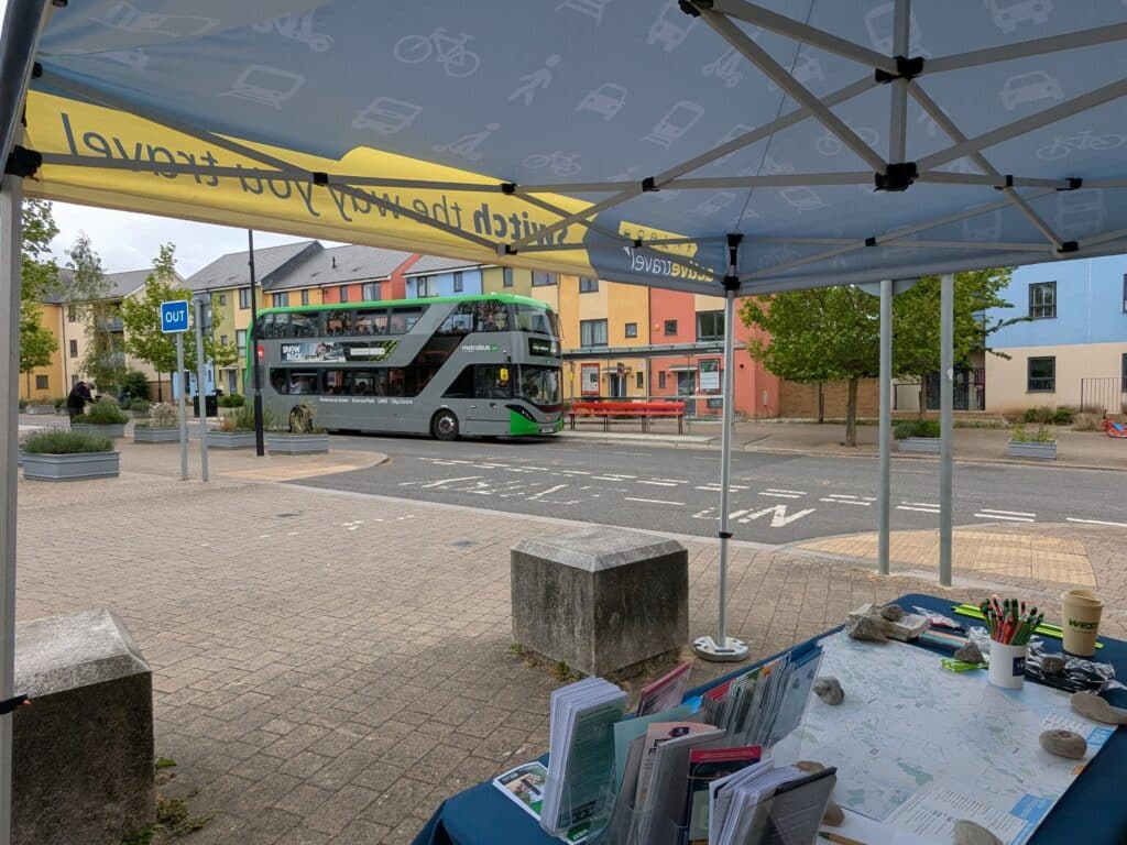 Photo of the view from under a pop-up gazebo looking out onto a residential street, with a metrobus stopped at a bus stop, colourful housing in the background, and a table with travel leaflets in the foreground.