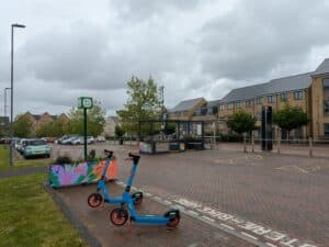 Photo of e scooters parked beside a shared mobility area with bike and scooter symbols, near a residential street with modern housing, parking spaces and a bus stop