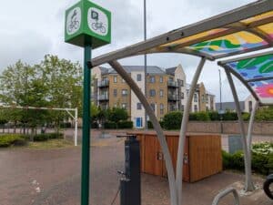 Photo of bicycle repair and parking point at a residential street, with a green sign showing bike parking symbols, sheltered by a colourful canopy near modern housing.