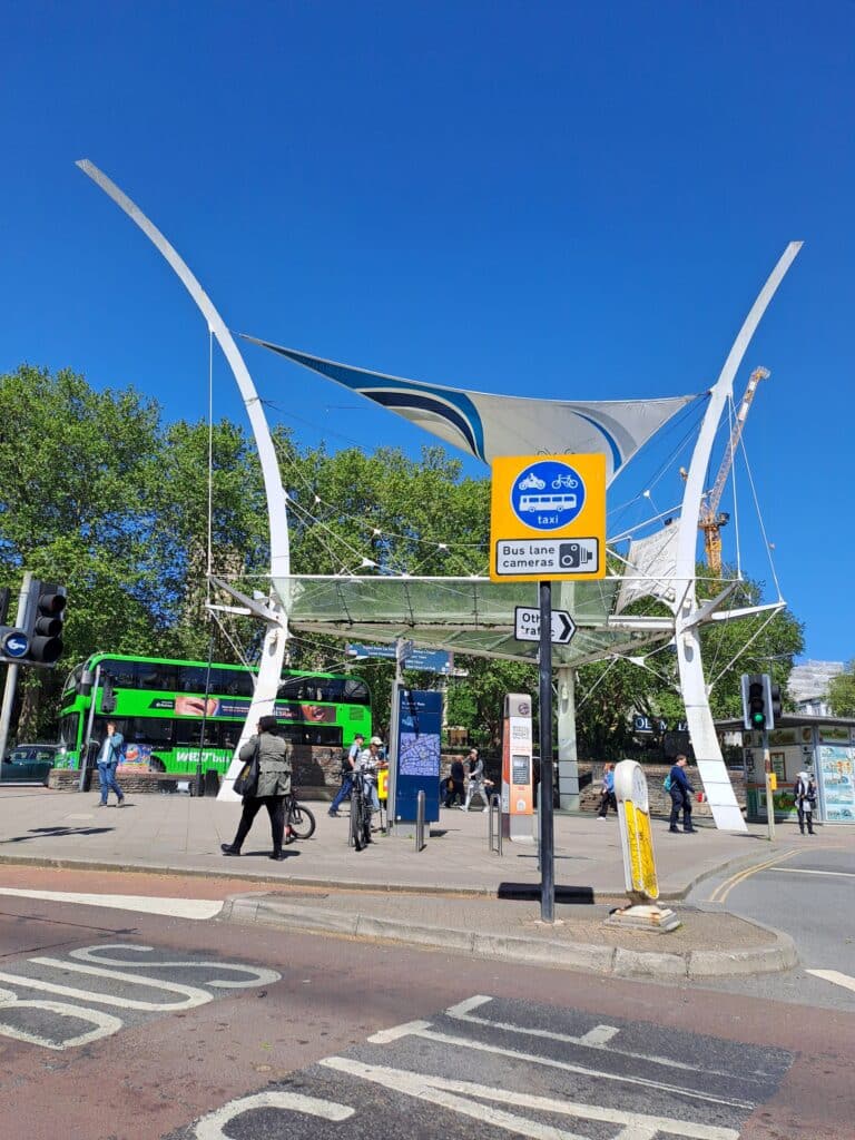 Modern white canopy structure, with a green double-decker bus in the background, and pedestrians under a clear blue sky.