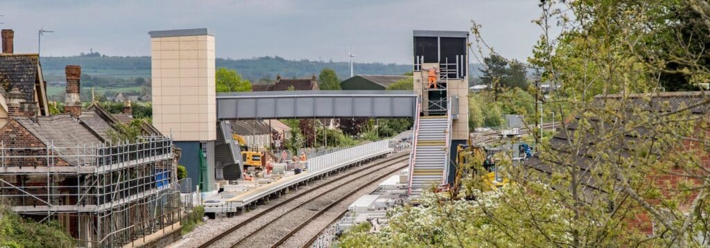 Charfield railway station under construction with a new pedestrian footbridge spanning multiple tracks. Workers in high-visibility clothing are on the platforms and stairs, with construction equipment nearby. Residential houses and greenery surround the station, and rolling countryside is visible in the background.