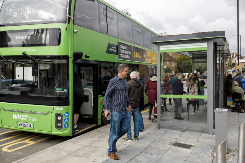 A bright green double-decker bus is stopped at a modern bus shelter while passengers board and wait nearby. Several people are standing around the stop, including a man walking in the foreground holding a phone. The bus displays “WESTbus” branding and appears to be part of a public transit service. The scene is outdoors on a cloudy day in what looks like a suburban or urban neighborhood street.