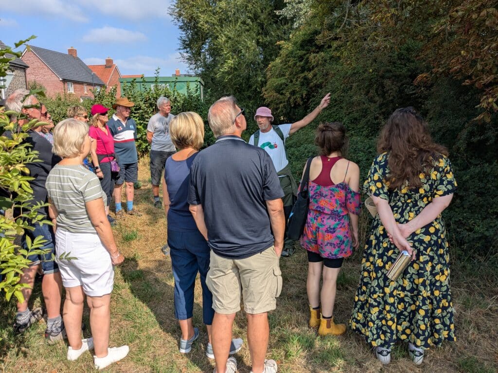 oto of a group of people gathered closely together on a grassy path beside a dense hedge and mature trees during an outdoor guided walk. The walk leader gestures towards a tree while speaking. Houses and garden vegetation are visible in the background, with bright sunshine and blue sky overhead.