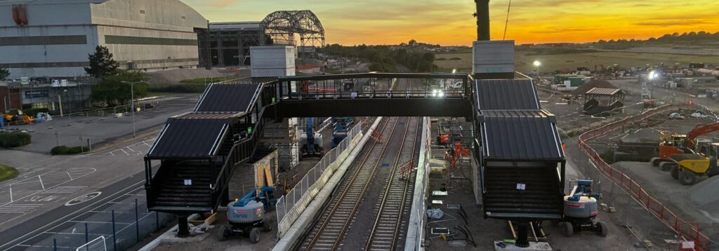 Railway station construction project featuring a new pedestrian footbridge over train tracks, surrounded by industrial facilities, construction equipment, and sunset lighting.