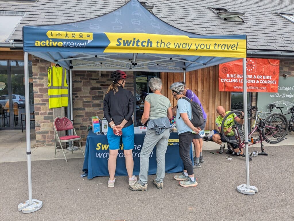 = Photo of people wearing cycle helmets gathered around an Active Travel information stand beneath a blue and yellow canopy displaying the slogan “Switch the way you travel.” The stand is set up outside a stone-fronted building, with leaflets and information on a table. To the right, a bicycle mechanic repairs an upturned bike. A high-visibility vest hangs from the tent frame, and several bicycles are parked nearby.