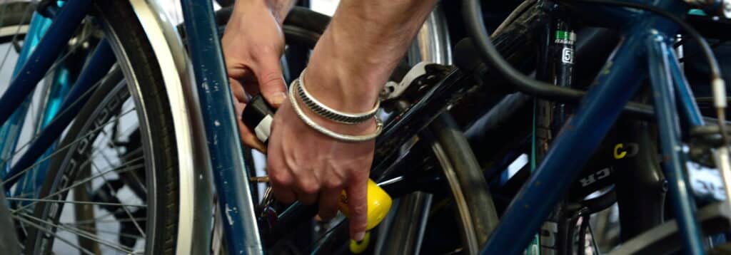 Close-up of a person securing a bicycle with a yellow lock in a cycle parking area. Several bicycles are parked closely together, with the focus on the person's hands fastening the lock around the bike frame.