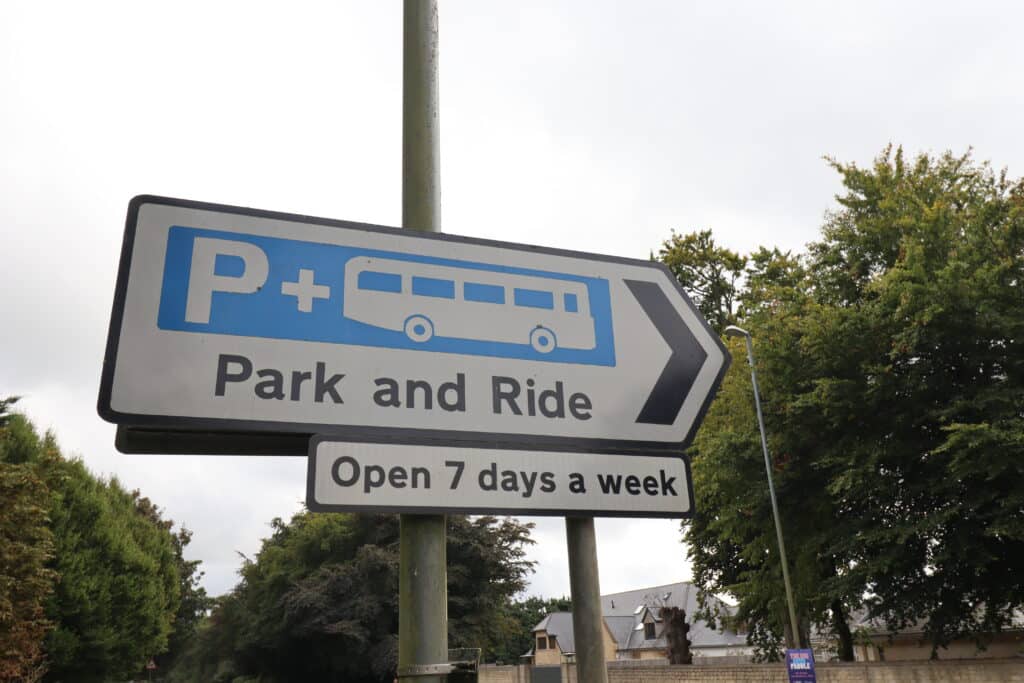 Roadside directional sign for a Park and Ride facility, featuring a blue parking and bus symbol with an arrow pointing right. A smaller sign below reads “Open 7 days a week”. Trees, lampposts and houses are visible in the background under an overcast sky.