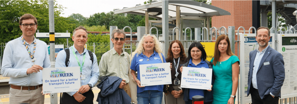 Various people stood in a row infront of Ashley Down Rail stations main platform, holding posters for the "Better Transport Week" campaign.