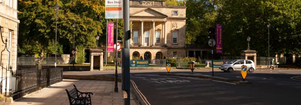 View along a wide city street with a bus stop in the foreground, facing the Holburne Museum. A bench and railings line the pavement, while a few cars travel on the road. Trees with autumn foliage frame the museum, with directional traffic islands and banners visible in the scene.