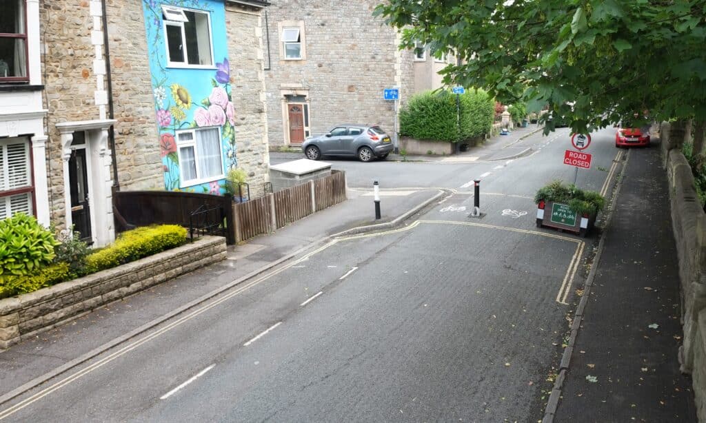 A residential street with a modal filter that allows people walking, wheeling and cycling to pass while restricting through traffic. Two black-and-white bollards and large planters create the filter, with cycle lane markings and signs indicating the road is closed to motor vehicles but open to cyclists. Stone terraced houses line the street, including one with a colourful floral mural, while mature trees overhang the pavement.