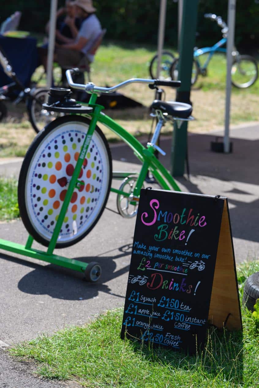 A bright green smoothie bike with a colourful front wheel display stands outdoors at a community event. In the foreground, a chalkboard sign advertises pedal-powered smoothies for £2 and a selection of hot and cold drinks. People sit under a gazebo in the background, with another bicycle visible behind them on a sunny day.
