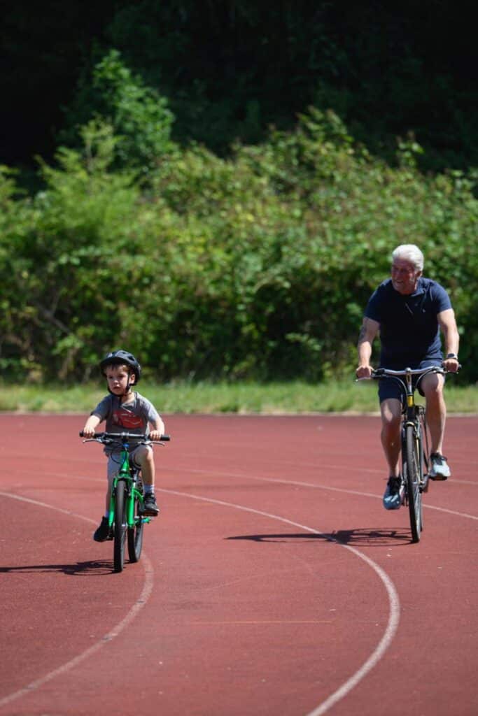 A young child wearing a cycle helmet rides a small green bicycle around an outdoor running track, while an older adult cycles behind them. Trees and greenery form the background on a bright, sunny day.